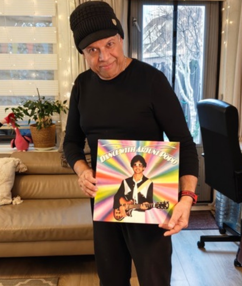Krishn Malhoe today, in a black cap and sweater, holding the Dance With Arjune Popo LP in his living room